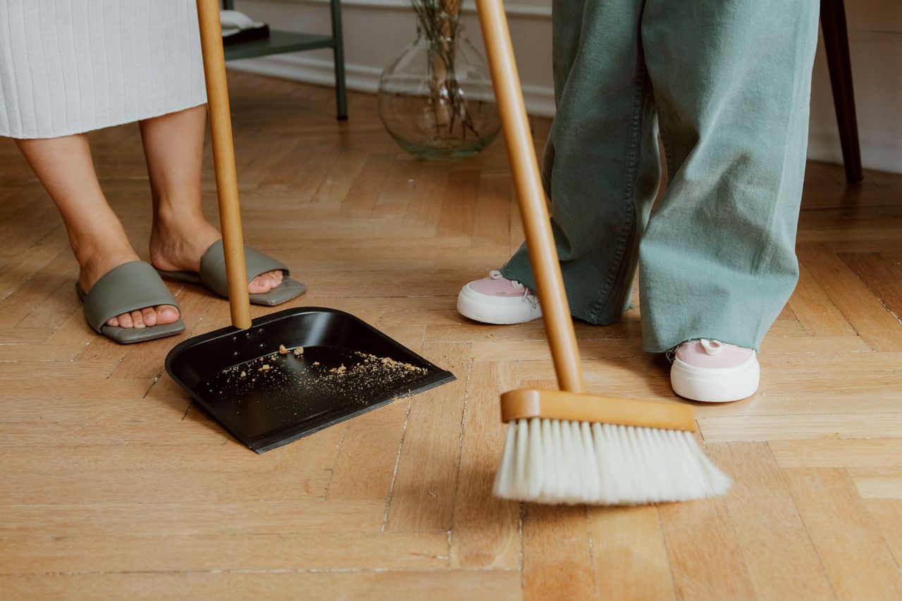 hero-img-02 Close-up of people cleaning wooden floor with broom and dustpan indoors.
